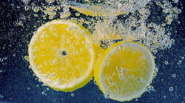 Rodaja de limón bajo el agua en agua de soda o limonada con burbujas Refrescante cóctel gaseoso tónico de soda Primer plano de limones y cubitos de hielo en vaso Cal en salpicaduras de agua con gas bebida fría