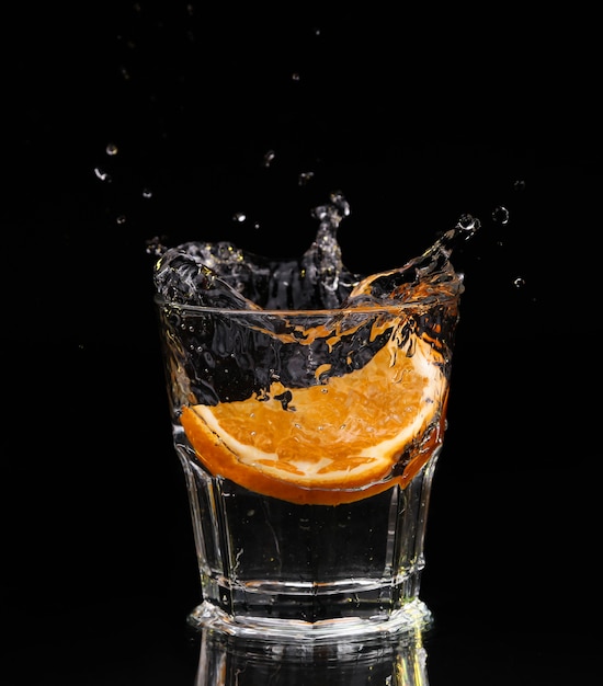 Rodaja de limón chapoteando en un vaso de agua con un chorro de gotas de agua en movimiento suspendido en el aire sobre el vaso sobre un fondo oscuro.
