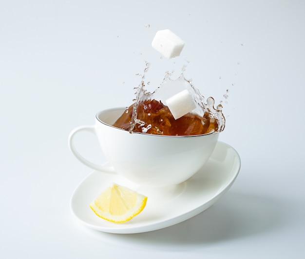 Rodaja de limón en platillo y cubos de azúcar cayendo en la taza de té con salpicaduras. Salpicaduras congeladas, levitación y equilibrio.