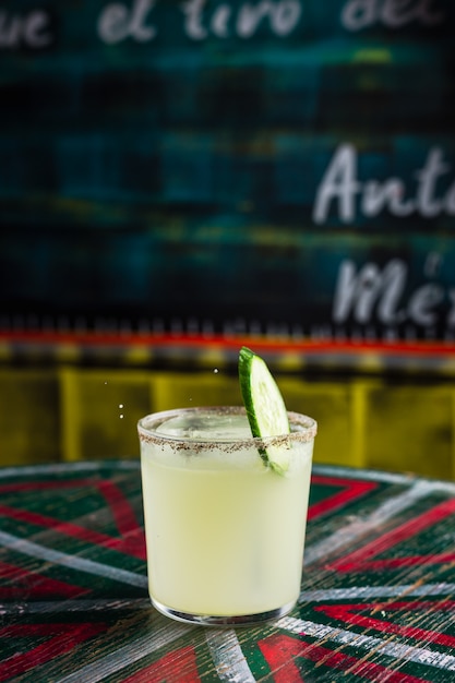 Una rodaja de pepino cayendo en un cóctel en un vaso lowball de rocas con un gran cubo de hielo, borde picante en el vaso