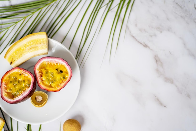 Rodajas de maracuyá, longan, plátano en un plato blanco. Frutas exóticas y hojas tropicales sobre fondo de mármol blanco con espacio para copiar su texto. Comida, concepto de verano. Vista superior.