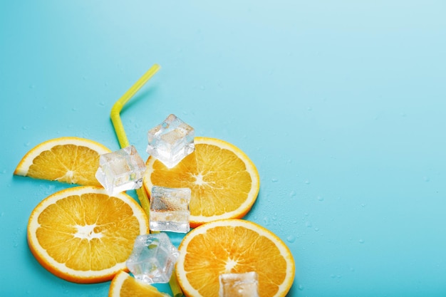 Rodajas de naranja y cubitos de hielo con una pajita sobre un fondo azul en forma de cóctel. El concepto de la bebida limonada es una composición refrescante de verano. Vista superior