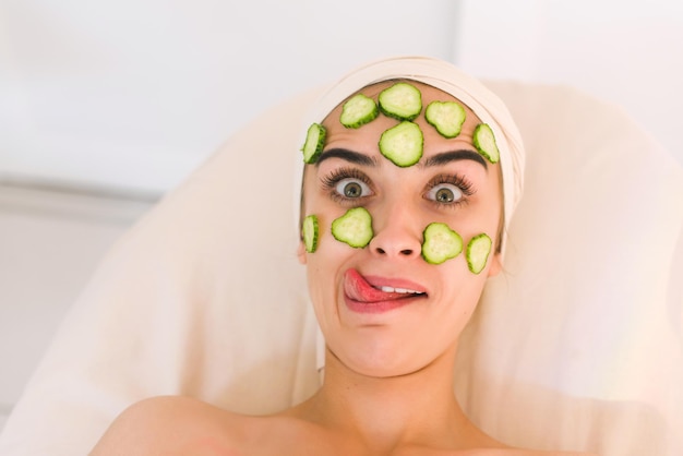 Rodajas de pepino en los ojos. Mujer joven con máscara facial de pepino en salón de belleza. La niña hace una mueca, muestra su lengua y entrecierra los ojos.