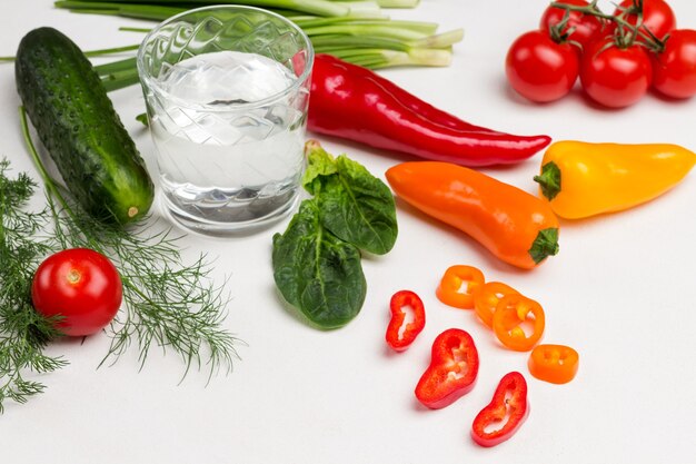 Rodajas de pimiento picado. Tomates y pimientos de colores. Vaso de agua, hojas de espinaca y eneldo y cebolleta. Fondo blanco. Vista superior