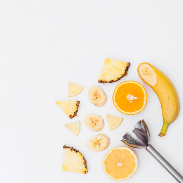 Foto rodajas de plátano; piña; mitades de naranjas con batidora de mano sobre fondo blanco