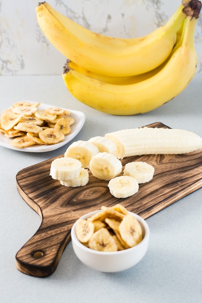 Rodajas de plátano en una tabla de cortar y chips de plátano en un recipiente sobre la mesa. Comida rápida. Vista vertical