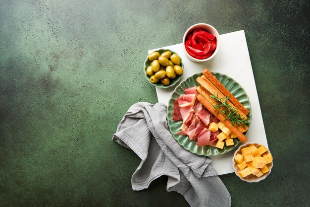 Rodajas de prosciutto o jamón palitos de grissini deliciosos con prosciutto queso romero aceitunas en plato verde en fondo oscuro aperitivos mesa con bocadillos italianos antipasto vista superior espacio de copia