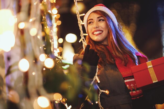 El rodaje de una joven alegre con una caja de regalo roja se está divirtiendo en la calle de la ciudad en Navidad.