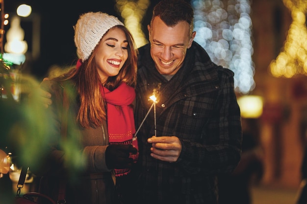 El rodaje de una pareja sonriente con bengalas está disfrutando en la víspera de Año Nuevo en la calle de la ciudad con luces navideñas.