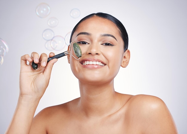 Rodillo facial de belleza y una mujer en estudio para dermatología de brillo de piel o cosmética natural Retrato de modelo femenino feliz para masaje facial y beneficios para el cuidado de la piel en burbujas y fondo blanco