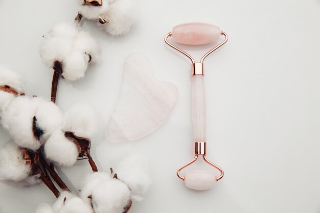 Rodillo facial de cristal de cuarzo rosa y herramienta de masaje jade Gua sha sobre una mesa blanca. Masaje facial anti-edad para un tratamiento lifting y tonificante natural en casa