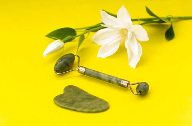 Rodillo facial de cuarzo verde cristal y herramienta de masaje jade Gua sha sobre piedras y flor blanca sobre fondo amarillo. Masaje facial antiedad para un lifting y tonificación natural en casa.