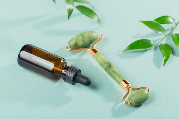 Rodillo facial de jade verde, tratamiento antiedad y hojas verdes sobre fondo azul claro. Herramienta de masaje para el cuidado de la piel facial, concepto de tratamiento de belleza y auto SPA.
