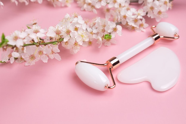 Rodillo facial de piedra gua sha para masaje facial sobre un fondo rosa con cereza floreciente Cuidado de la piel facial cosmético y spa Concepto de tratamiento natural