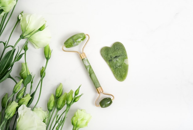 Rodillo de jade para masaje facial. Herramientas de masajeador facial gua sha verde sobre el fondo blanco de mármol con espacio de copia plano. Tratamiento antiedad, lifting y tonificante, acupresión. Flores de Eustoma