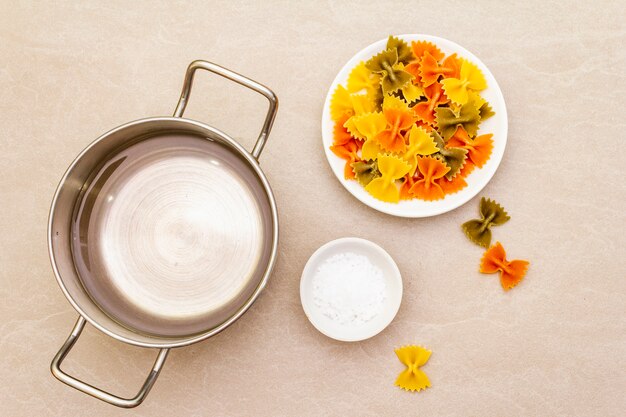 Rohe Farfalle mit Wasser in Topf und Salz. Mehrfarbige Teigwaren in der weißen Platte auf Steinoberfläche. Für Kinder sich vorbereiten, Draufsicht, Abschluss oben