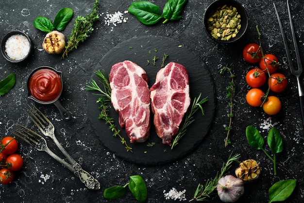 Rohe frische Schweinesteaks mit Gewürzen und Kräutern Draufsicht auf schwarzem Steinhintergrund