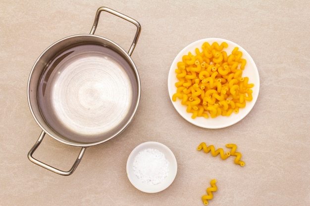 Rohe Fuzilli mit Wasser in Topf und Salz. Traditionelle italienische Teigwaren in der weißen Platte auf Steinoberfläche. Vorbereitung für Kinder, Ansicht von oben