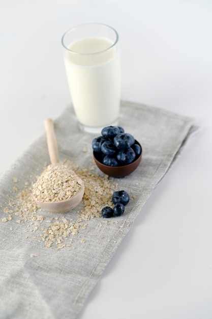 Rohe Haferflocken mit schwarzen Blaubeeren und einem Glas Milch Hoher Winkel frischer Blaubeeren und ein Holzlöffel mit Haferflocken auf dem Tisch neben einem Glas Milch und auf weißem Hintergrund im hellen Raum