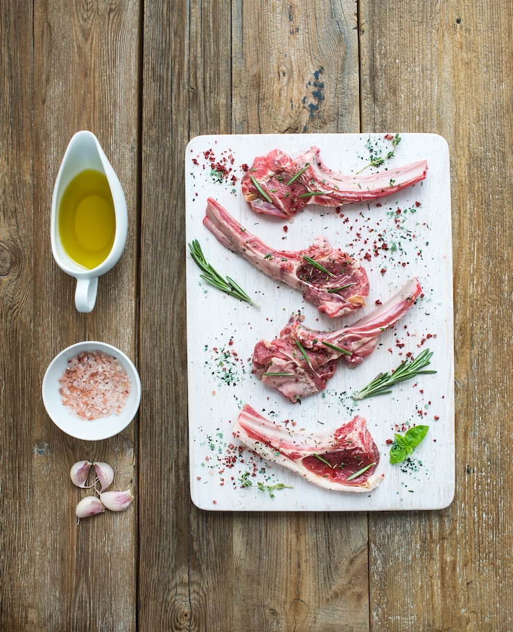 Foto rohe lammkoteletts. lammkarree mit knoblauch, rosmarin und gewürzen auf weißem hackendem brett, öl in einer untertasse, salz, essgeschirr über rustikalem hölzernem hintergrund.