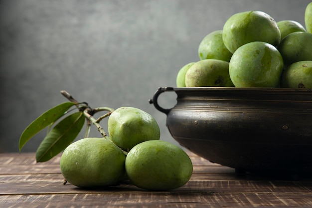 Rohe Mango, die traditionell in einem Messinggefäß angeordnet ist, und ein Haufen grüner Mango, die in der Nähe auf einer hölzernen strukturierten Basis mit selektivem Fokus auf grauem Hintergrund platziert sind