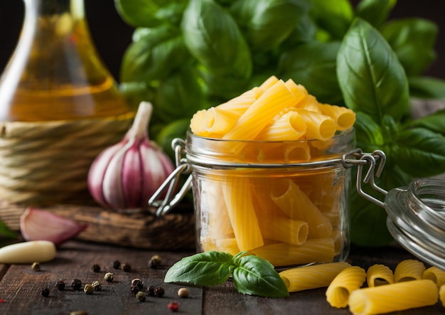 Rohe Penne-Nudeln im Glas mit Öl und Knoblauch-Basilikum-Pflanze auf Holztischhintergrund Makro