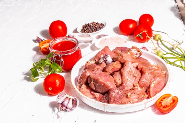 Rohe Schweinefleischstücke, Tomaten, Gewürze und Kräuter. Zutaten zum Kochen von gesundem Essen auf weißem Kitthintergrund. Kulinarisches Prozesskonzept, trendiges hartes Licht, dunkler Schatten, Kopierraum