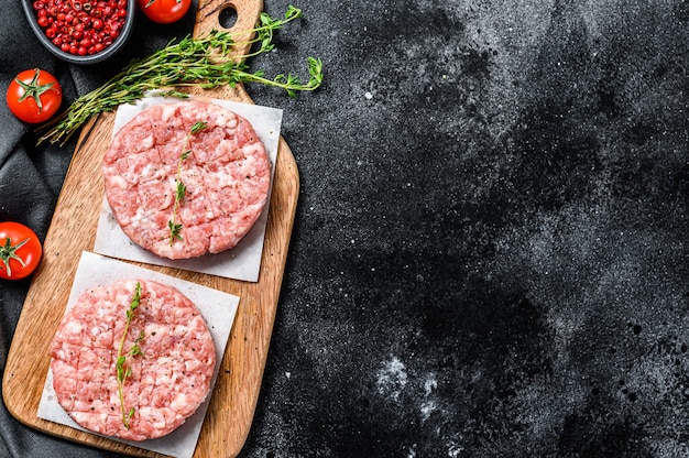 Rohe Schweinekoteletts, Hackfleischpastetchen auf einem Schneidebrett. Bio-Hackfleisch. Schwarzer Hintergrund. Draufsicht. Speicherplatz kopieren.