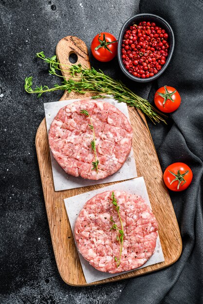 Rohe Schweinekoteletts, Hackfleischpastetchen auf einem Schneidebrett. Bio-Hackfleisch. Schwarzer Hintergrund. Draufsicht