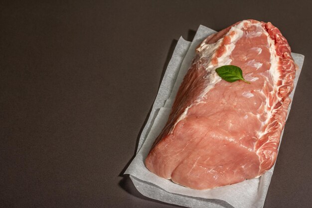 Rohe Schweinelende mit Basilikumblatt. Großes Stück Fleisch, Betonhintergrund aus schwarzem Stein. Trendiges hartes Licht, dunkler Schatten, Kopierraum