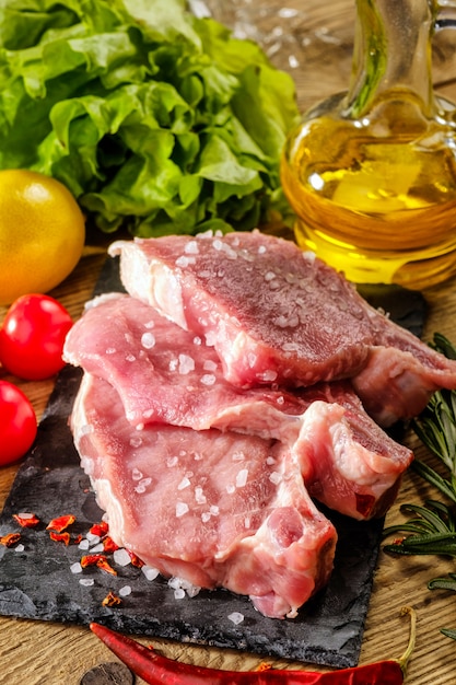 Rohe Schweinesteaks auf Steinbrett mit Kräutern, Tomaten und Zitrone.