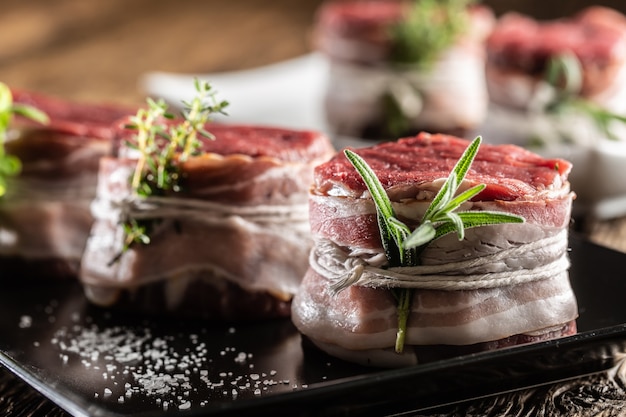 Rohe Steaks umwickelt mit Speck und frischen Kräutern auf einem Teller und einer Vintage-Holzoberfläche.