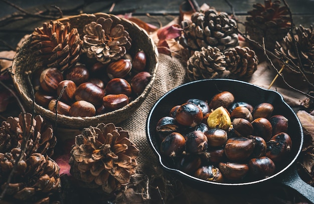 Rohe und geröstete kastanien in einer kleinen eisenpfanne und einer schüssel auf einem tisch mit herbstlaub und tannenzapfen