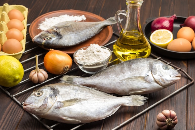 Roher Fisch mit Gemüse auf Grillrost. Ei und Mehl, Flasche mit Öl.