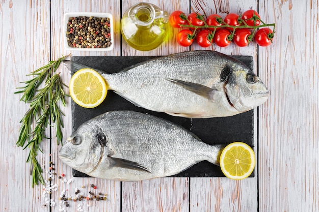 Roher Fischdorado auf schwarzem Schiefer-Schneidebrett und weißem Holzhintergrund mit Gewürzen, Tomaten, Rosmarin, Olivenöl und Zitrone. Ansicht von oben, flach mit Kopienraum für Text