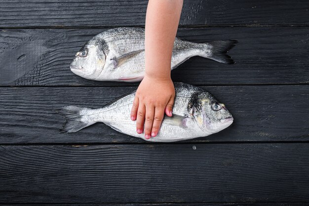 Roher ganzer Dorado oder Seebrassenfisch mit Kinderhand darüber auf schwarzem Holztisch