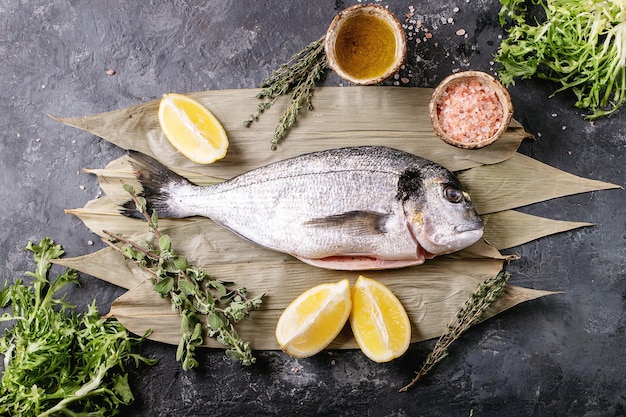 Roher ungekochter, frisch ausgenommener Seebrassen- oder Dorado-Fisch, serviert mit Zitrone, grünem Salat, Rosmarin, Olivenöl und rosa Salz auf Bambusblättern über einer dunklen Texturoberfläche. Flache Lage, Kopierraum.