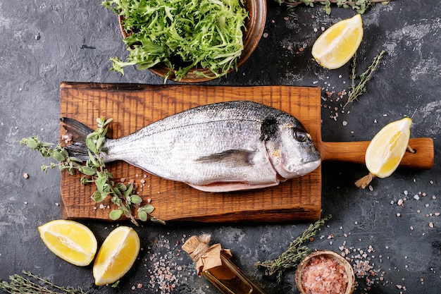 Roher ungekochter, frisch entkernter Seebrassen- oder Dorado-Fisch, serviert mit Zitrone, grünem Salat, Rosmarin, Olivenöl und rosa Salz auf einem Holzschneidebrett über einer dunklen Texturoberfläche. Flache Lage, Kopierraum.