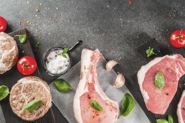 Rohes Bio-Fleisch. Auswahl verschiedener roter Fleischsorten: Schweineschnitzel mit Knochen, Schweinesteaks und Hühnerschnitzel in Schweinefettgitter. Mit Zutaten zum Kochen, grauer Steintisch