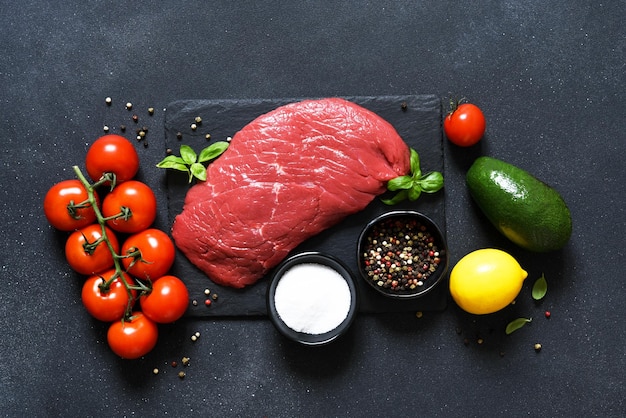 Rohes Fleisch mit Gewürzen und Gemüse: Avocado, Zitrone, Knoblauch und Tomaten auf einem Steinbrett auf schwarzem Hintergrund. Sicht von oben.