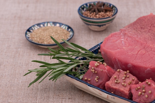 Rohes Fleisch mit Gewürzen und Kräutern in einem quadratischen Teller auf einem strukturierten Gewebe