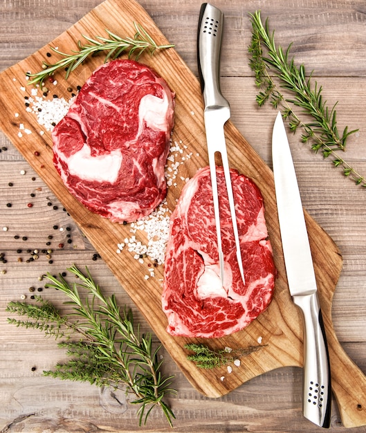 Rohes Frischfleisch Ribeye Steak mit Kräutern und Gewürzen auf Holzschreibtisch. Gabel und Messer zur Essenszubereitung