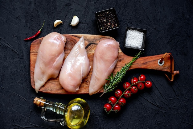 Rohes Huhn auf einem Holzschneidebrett mit Zutaten auf schwarzem Hintergrund.