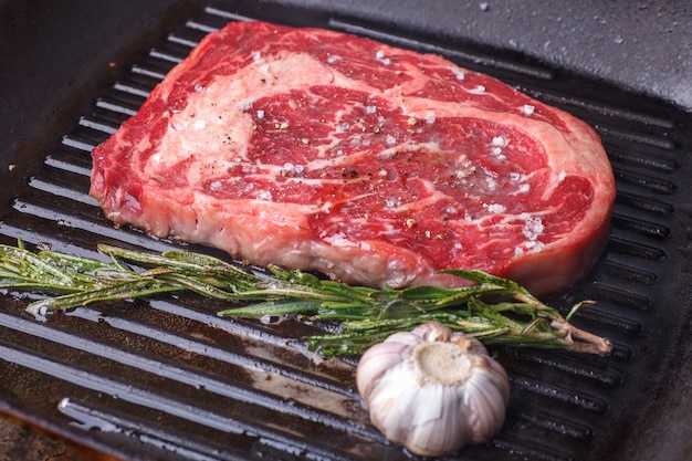 Rohes Marmor-Rindersteak, das in einer schwarzen Grillpfanne mit Knoblauch und Rosmarin geröstet wird