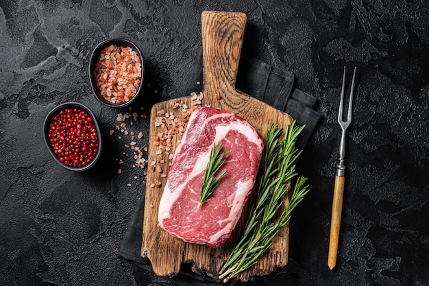 Rohes Rib-Eye-Steak, marmoriertes Rindfleisch auf Holzbrett mit Rosmarin. Schwarzer Hintergrund. Ansicht von oben.