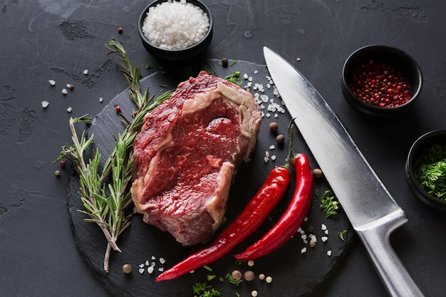 Rohes Rib-Eye-Steak mit Kräutern und Gewürzen. Kochzutaten für Restaurantgericht. Frisches Fleisch mit Chili und Messer auf Teller auf schwarzem Hintergrund.