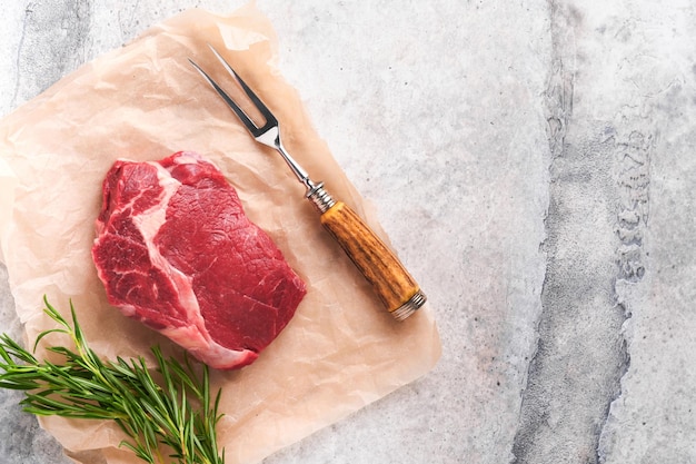 Rohes Rindersteak Frisches Rinder-Rib-Eye-Steak mit Gabel Rosmarin Salz und Pfeffer auf einem Stück Pergamentpapier auf grauem Steinhintergrund Draufsicht Mockup für Designidee