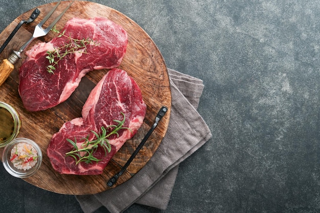 Rohes Rindersteak Marmoriertes rohes frisches Ribeye-Steak mit Rosmarinsalz und Pfeffer auf Schneidebrett auf dunklem Betongrund Rohes Rindersteak und Gewürze zum Kochen Mock up