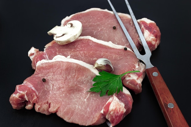 Rohes Rindfleisch mit Petersilie Knoblauch und Champignons und Fleischgabel auf dunklem Hintergrund Filetsteak Rindfleisch Frisches Fleisch in Reihe mit Gewürzen und Zutaten zum Kochen