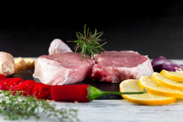 Rohes Schweinefleisch auf schwarzer Schieferplatte mit Gewürzbestandteil - Rosmarin, Ingwer, Paprikapfeffer, Zwiebel.